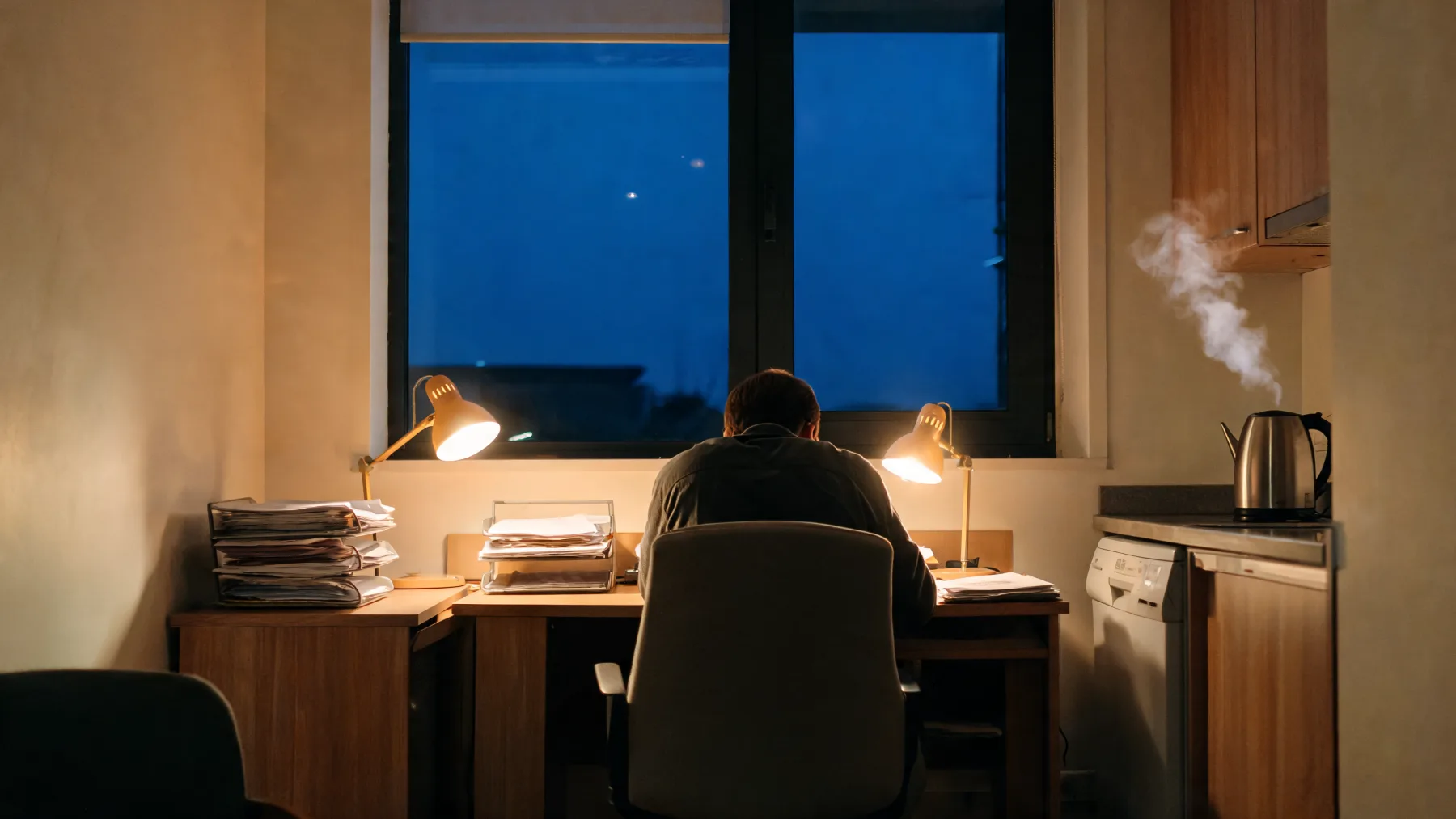 A small British office at dusk, desk lamps on against blue evening light.